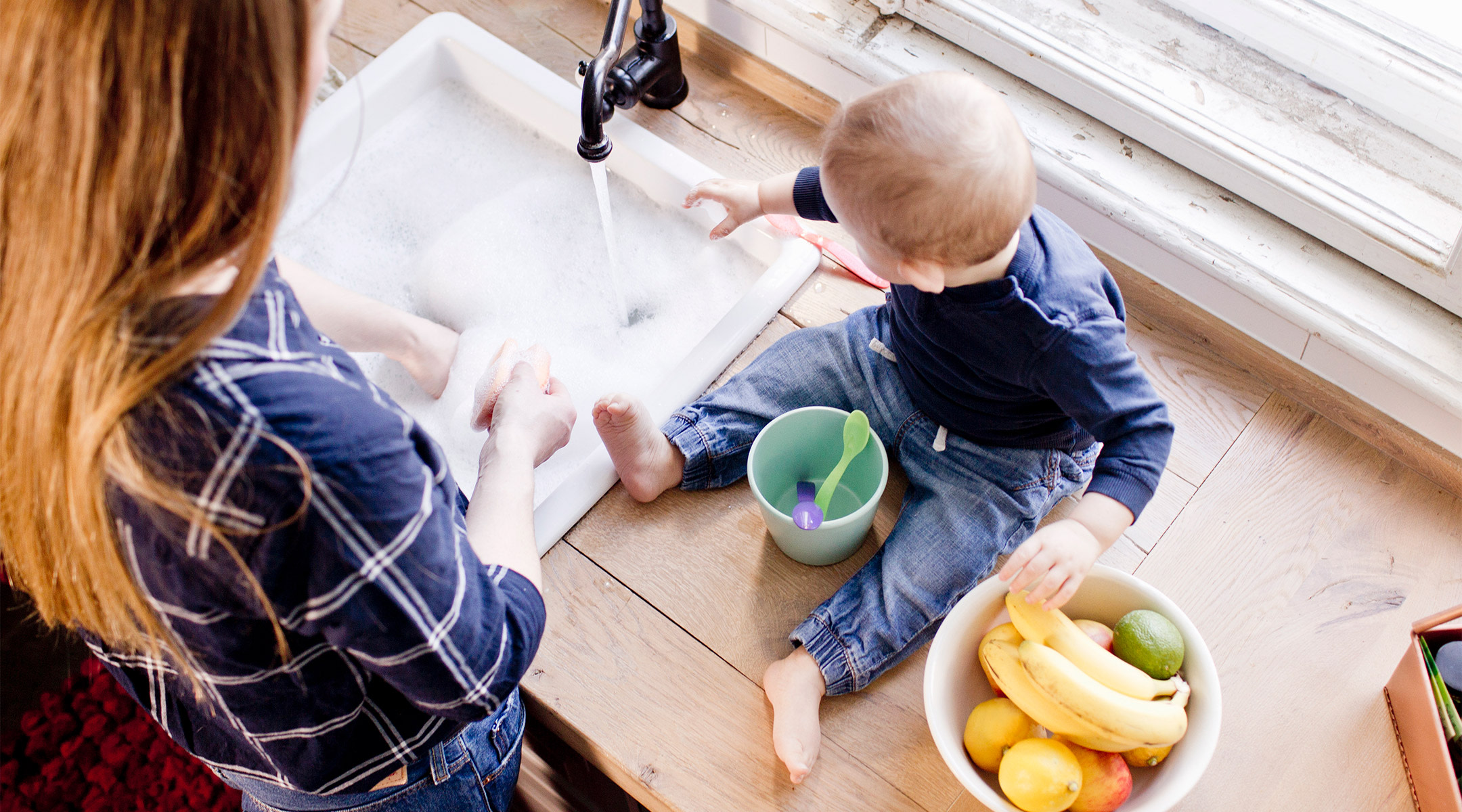 Mom doing dishes in the sink with baby sitting nearby