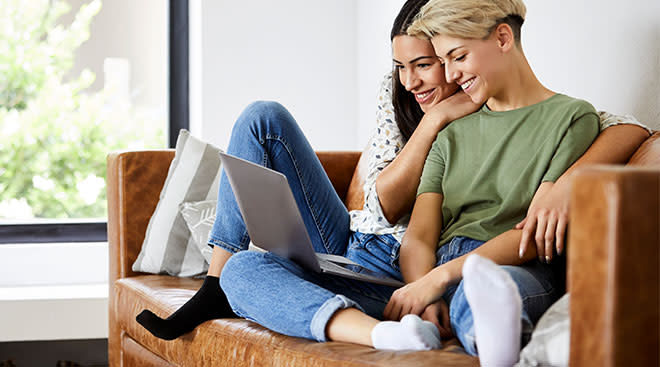 Same sex couple smiling and looking at laptop together on the couch. 