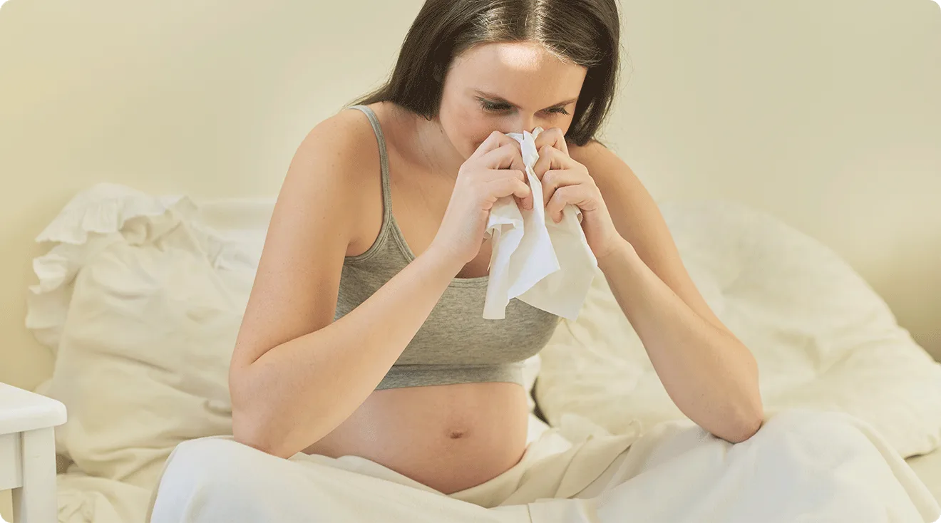 pregnant woman sick with a cold blowing nose into tissue at home