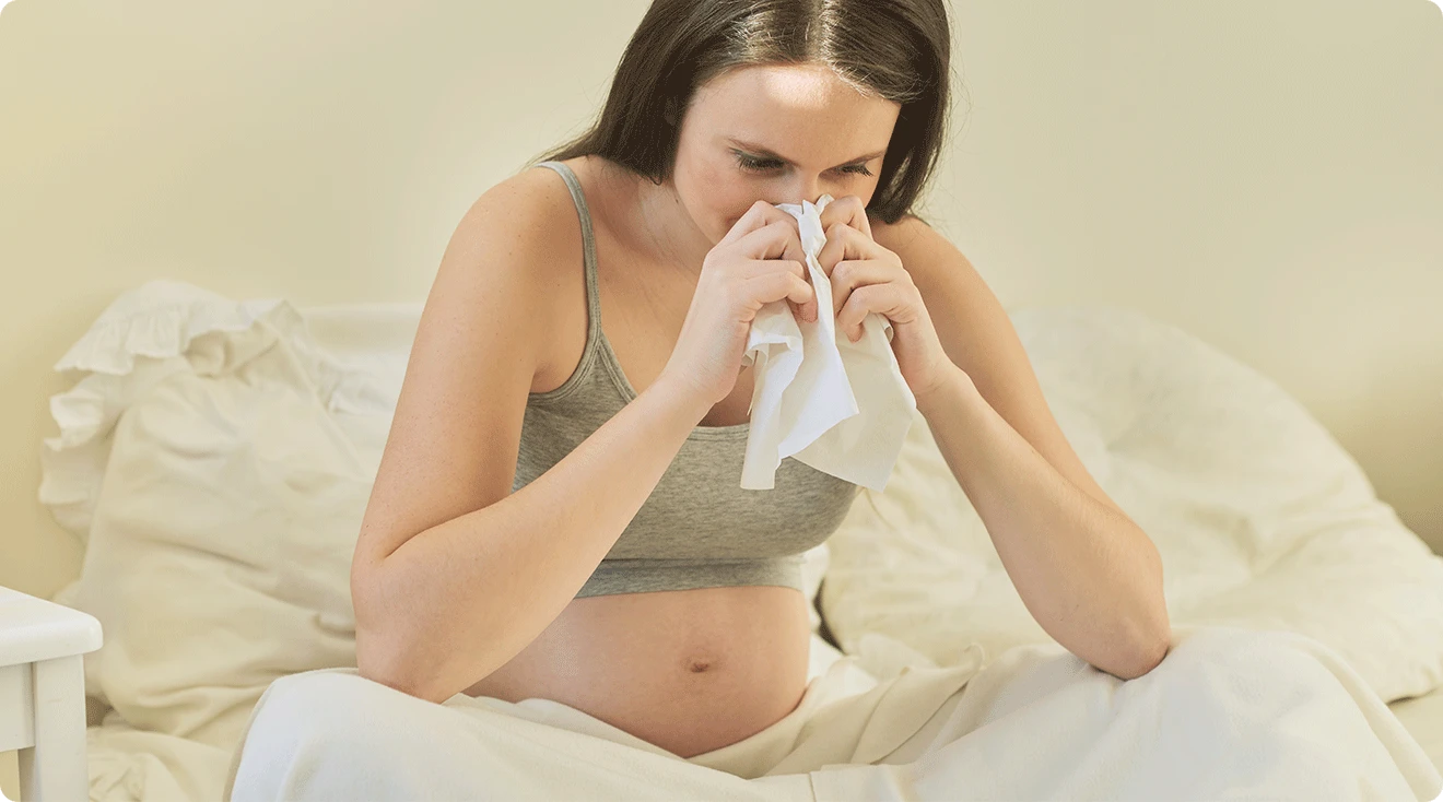 pregnant woman sick with a cold blowing nose into tissue at home