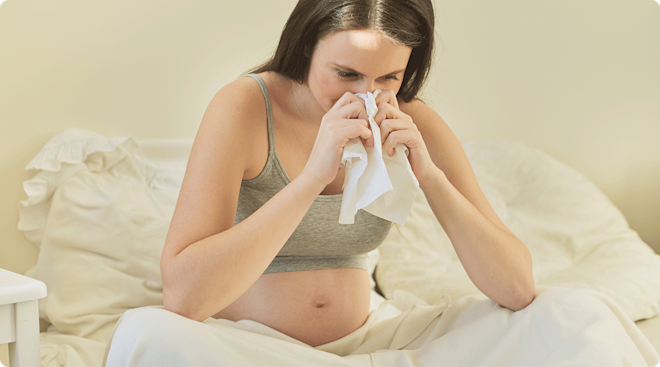 pregnant woman sick with a cold blowing nose into tissue at home