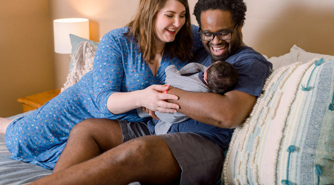 new dad with wife holding newborn baby at home on their bed