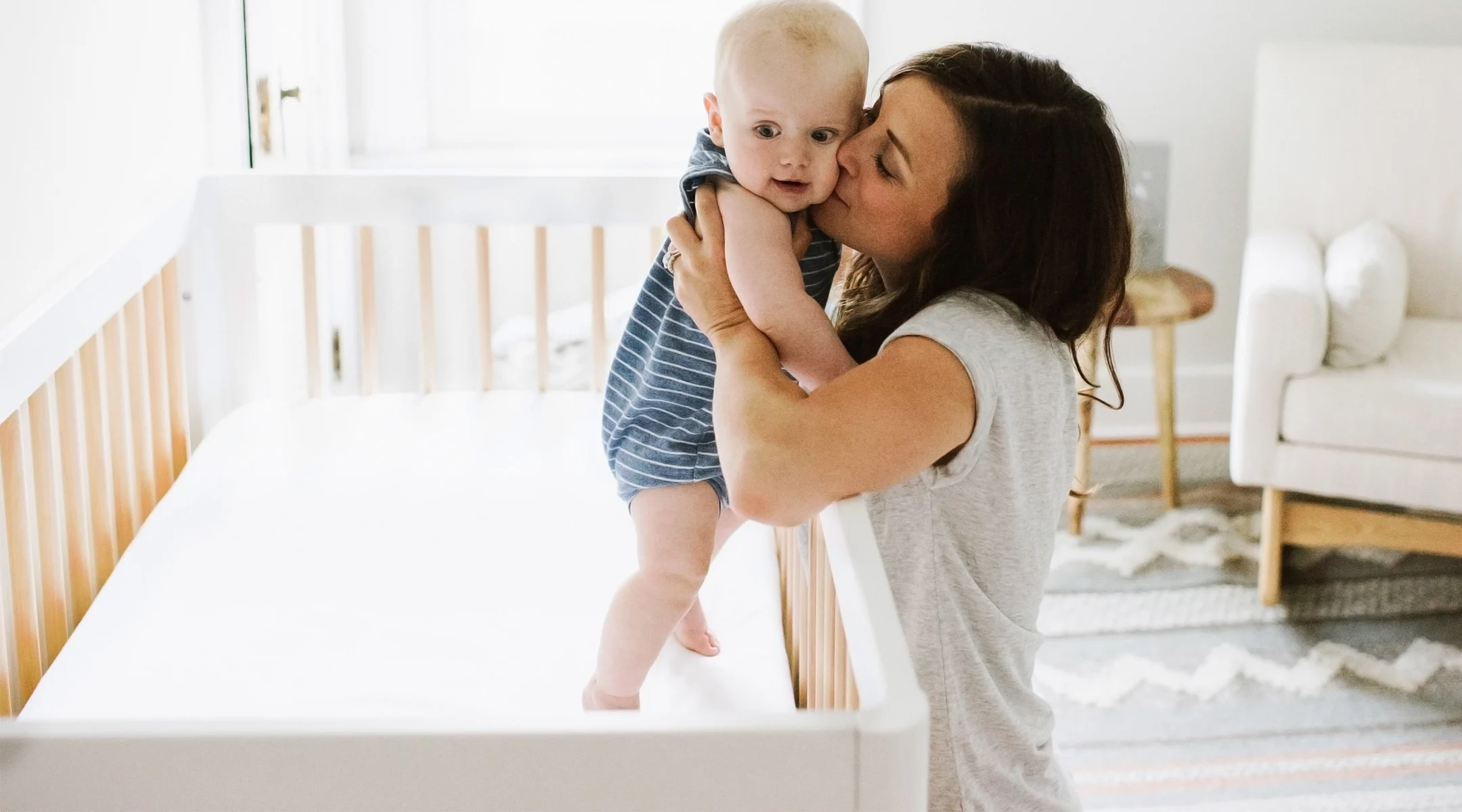 mom holds her baby by the crib and gives baby a kiss