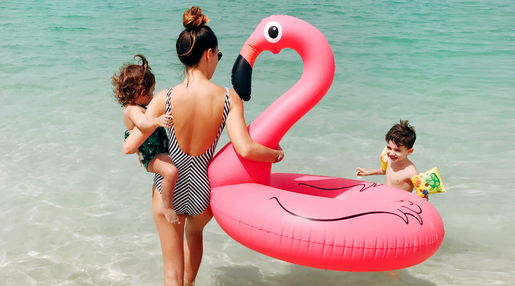 mom at beach traveling with her two young kids and holding a flamingo float