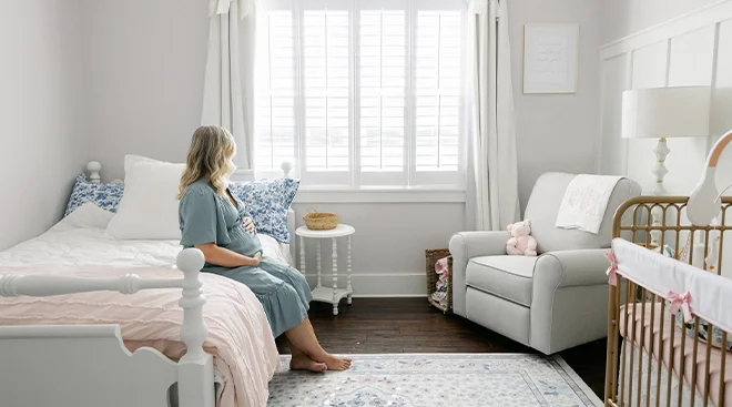 pregnant woman sitting on bed in baby's nursery room looking out window