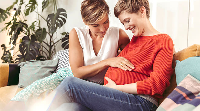 smiling same sex female couple sitting together and looking at one of their pregnant bumps