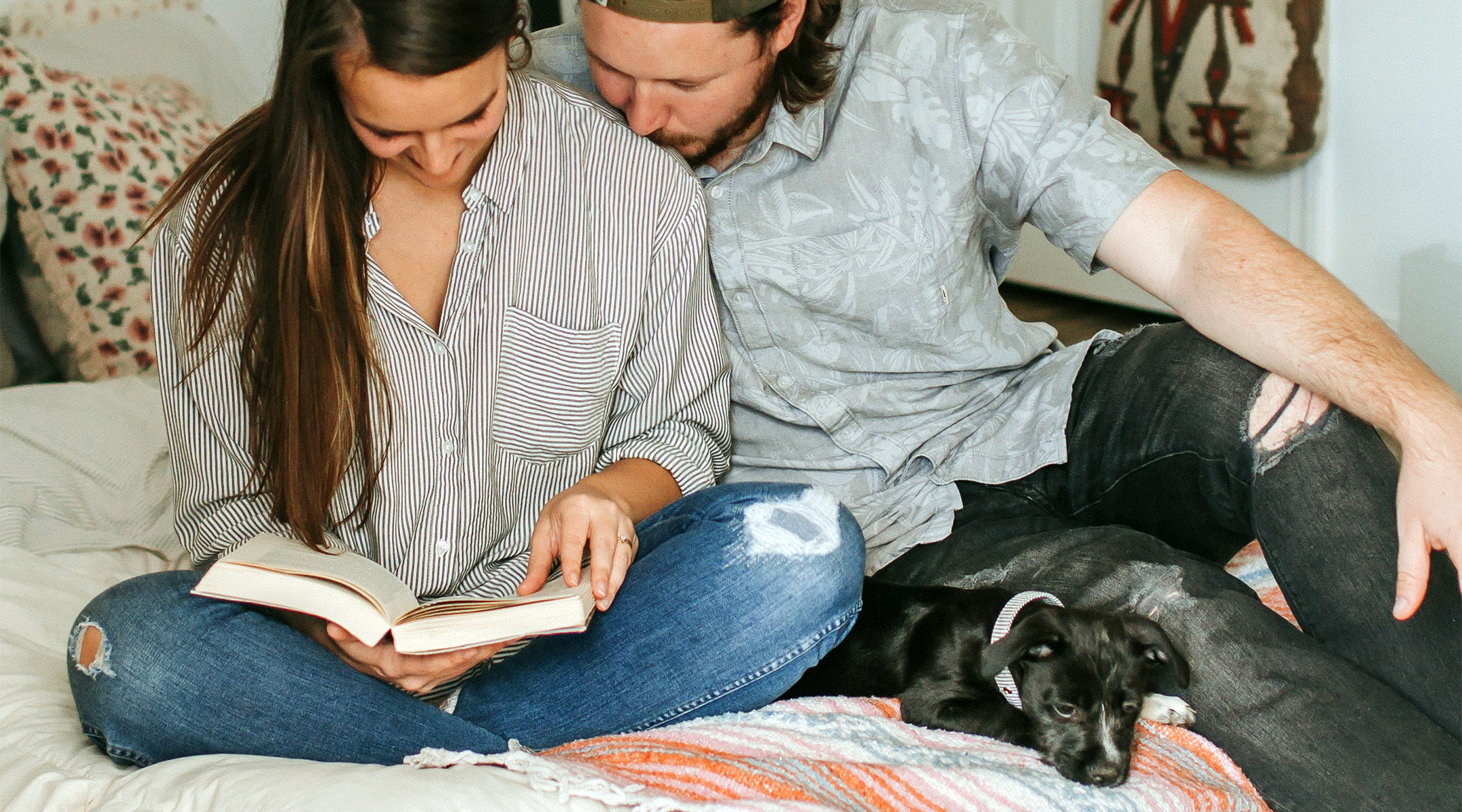 couple sitting on bed together reading a book with their puppy