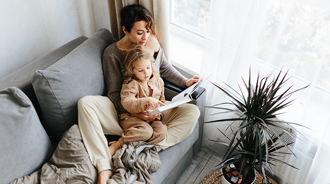 mother and toddler daughter sitting on the couch reading a book together at home