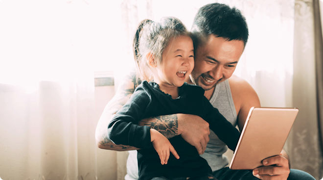 A father and young daughter laughing while looking at a tablet.
