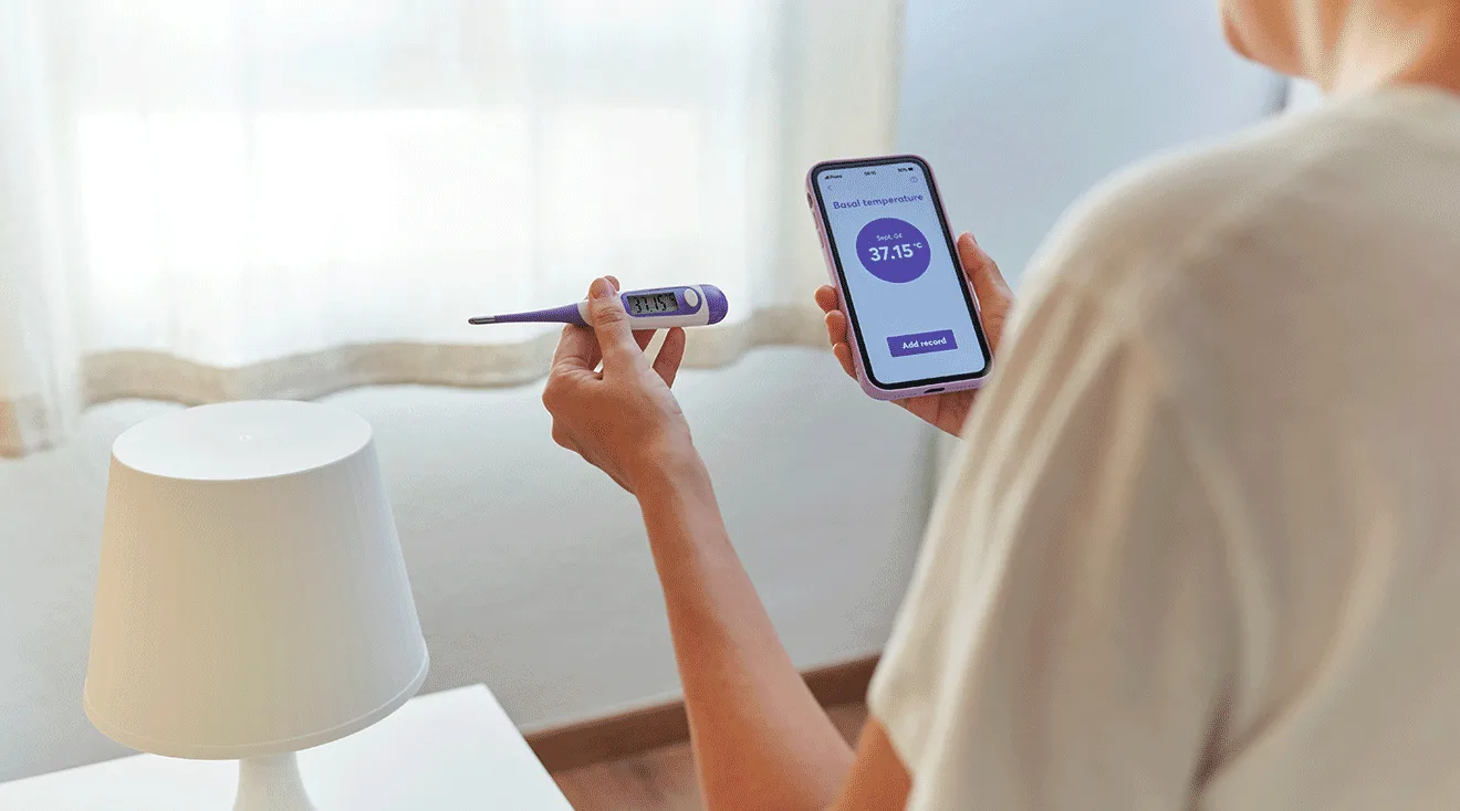 woman checking basal body temperature with thermometer and phone app