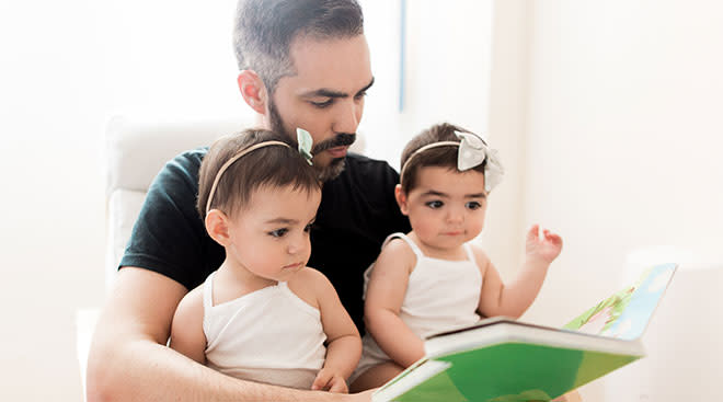 twin baby girls sitting on their dad's lap while he reads them a book