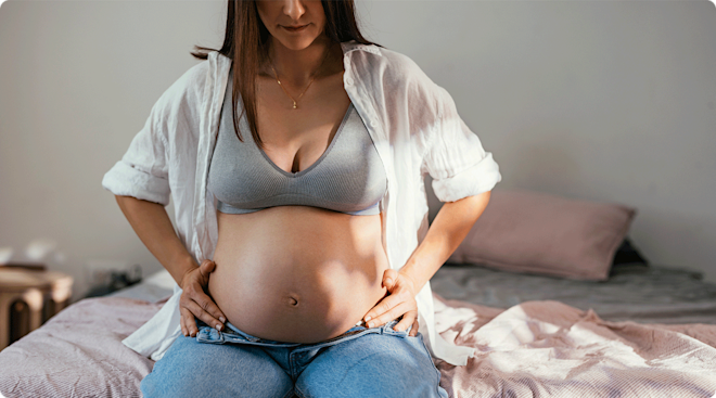 A pregnant woman sitting on the edge of her bed at home.
