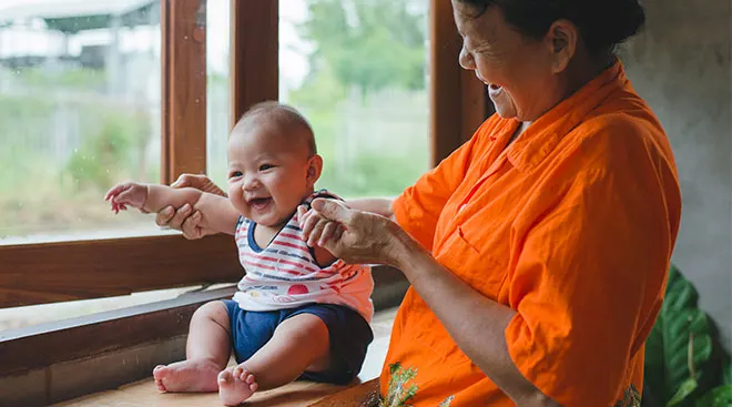 Grandma happily interacting with her baby grandson, holding his arms up while he laughs.