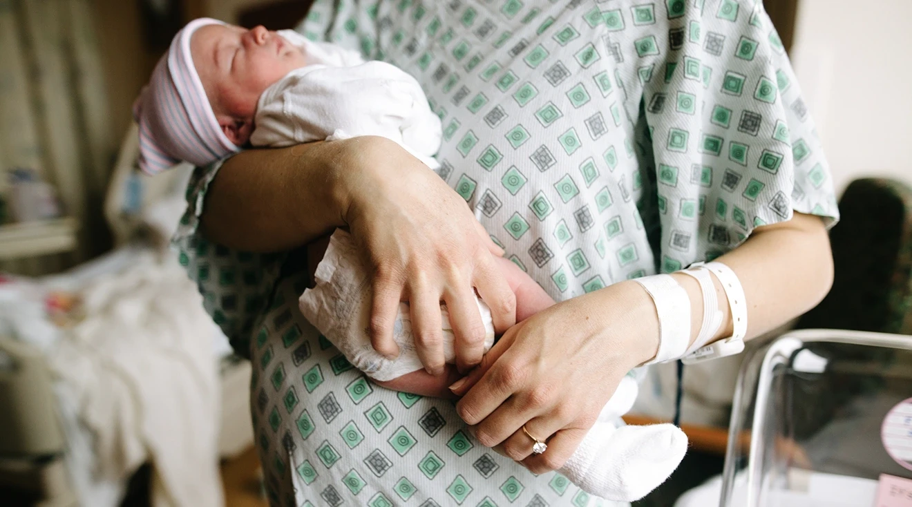 mother wearing hospital gown and holding newborn baby in hospital room after labor and delivery