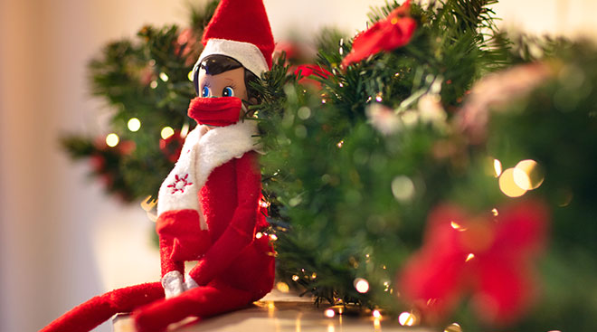christmas elf on the shelf sitting by greenery and wearing a mask