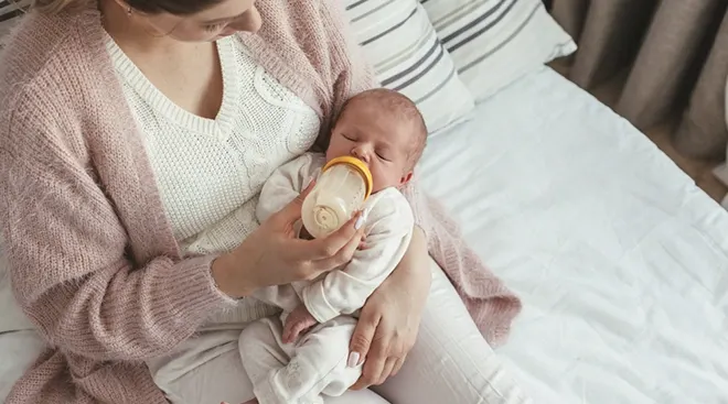 mother feeding newborn baby a bottle while sitting on bed at home