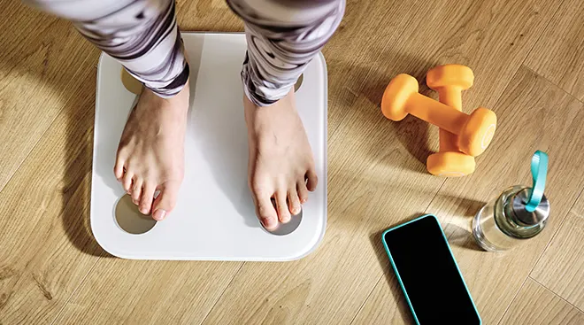 overhead view of woman's feet on digital scale next to smartphone and dumbbell weights