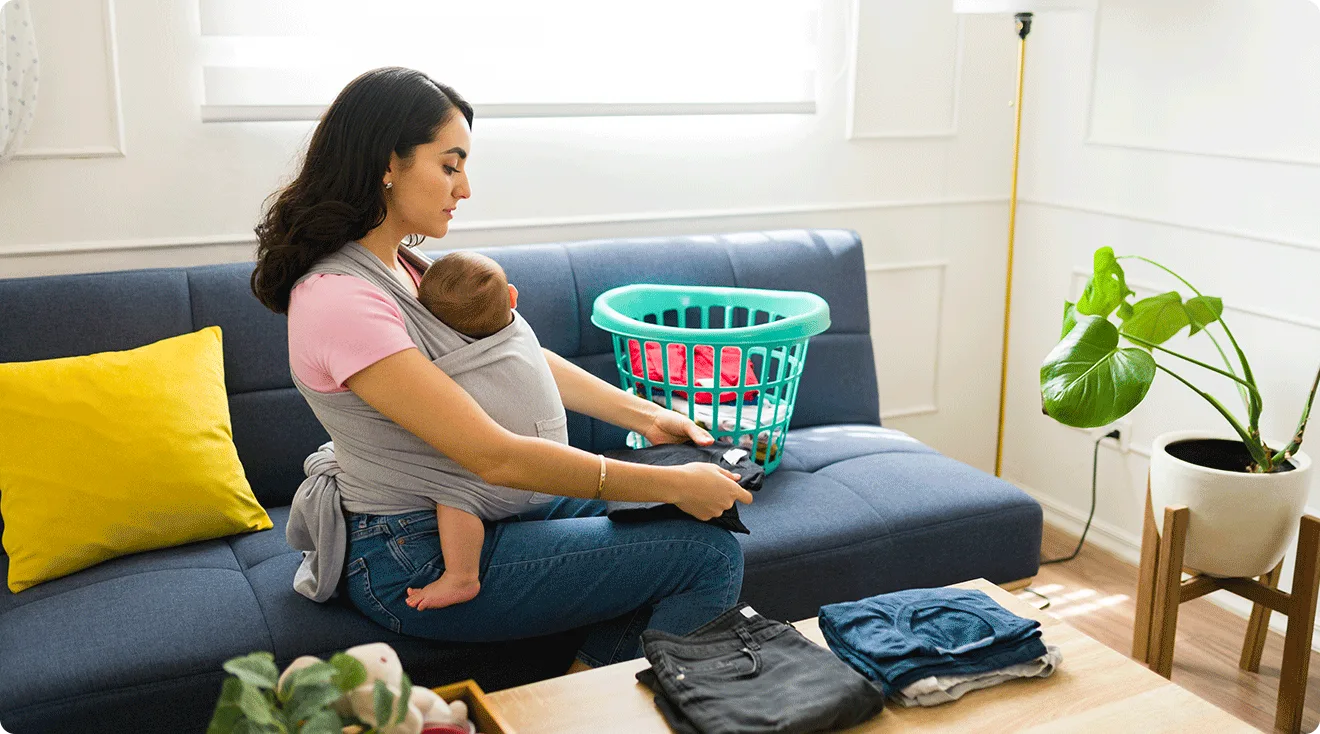 mom holding baby in baby carrier while folding laundry at home