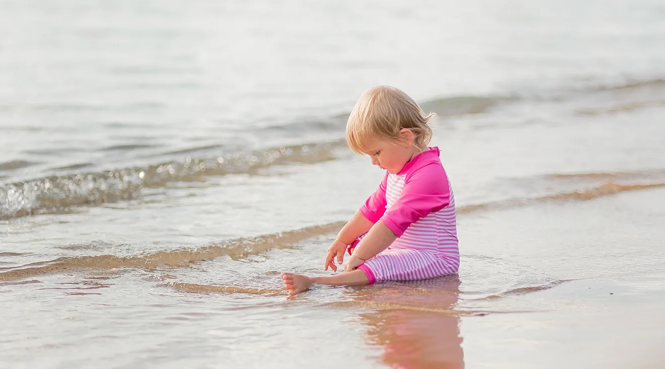toddler sitting by the ocean wearing bright pink swimsuit