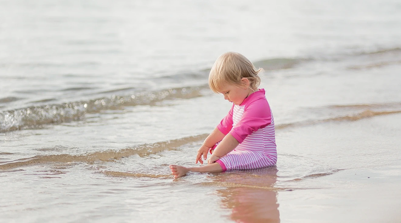toddler sitting by the ocean wearing bright pink swimsuit