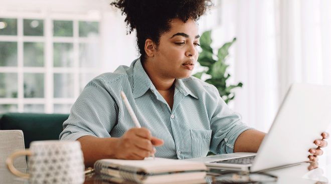 woman writing and looking at laptop while sitting at table at home