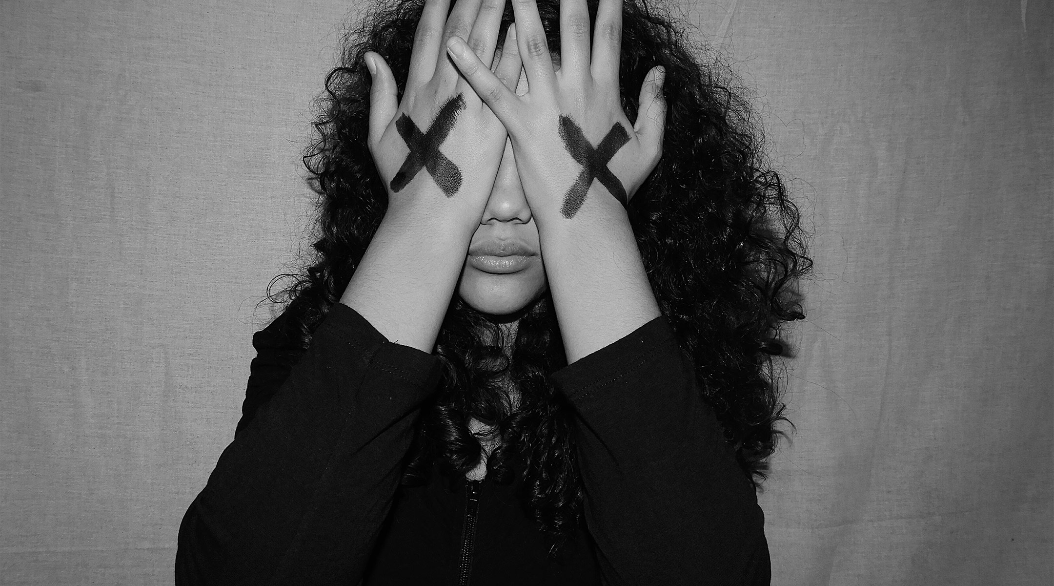 upset woman covering her face with her hands. Her hands have x's drawn on them.