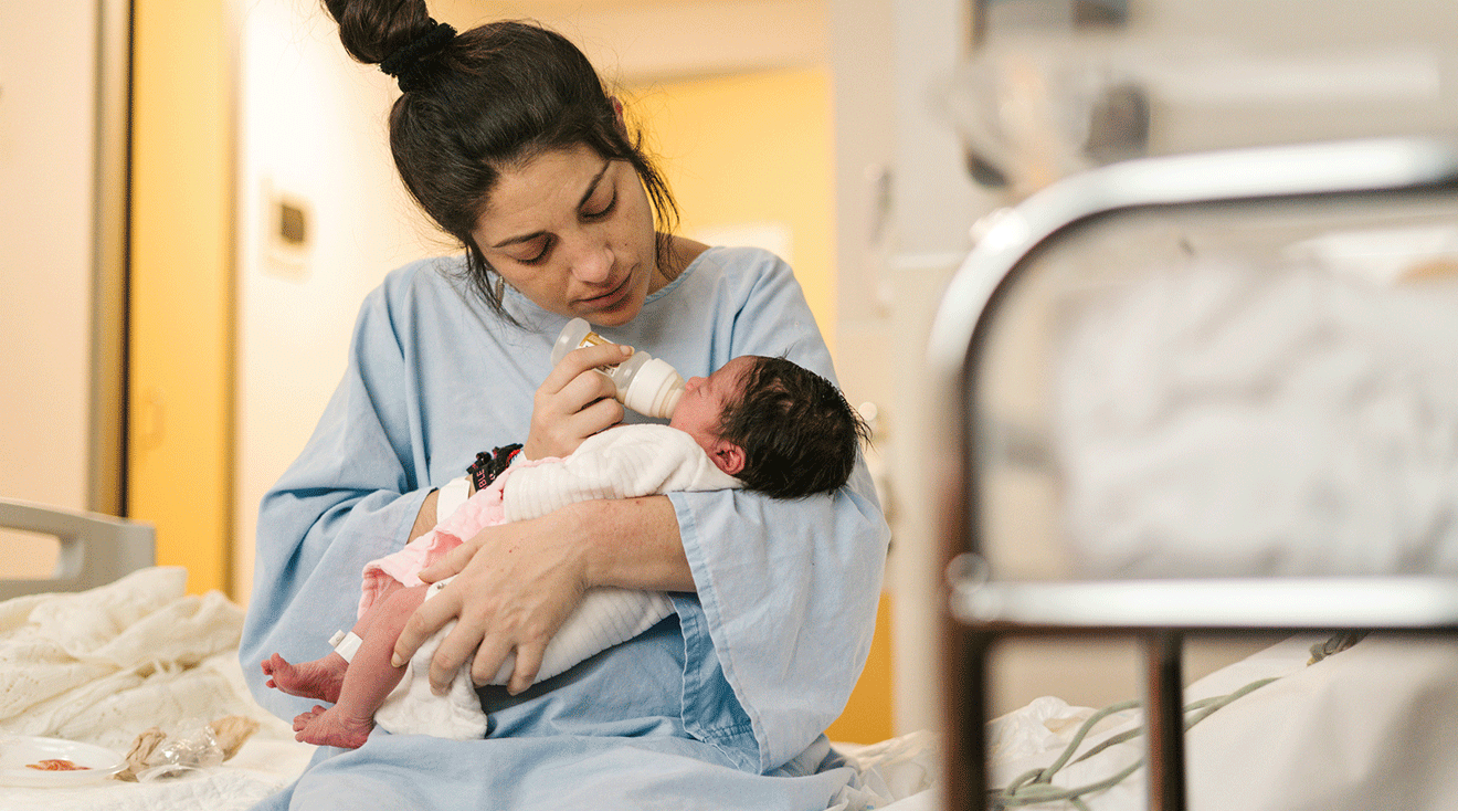 mother giving newborn baby a bottle while sitting on hospital bed after labor and delivery