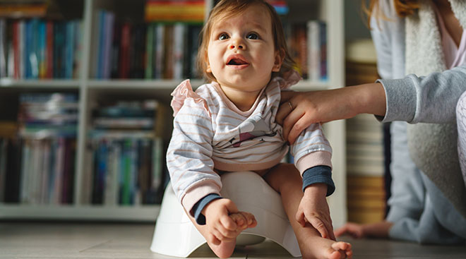 young baby sitting on small potty at home with help from mom