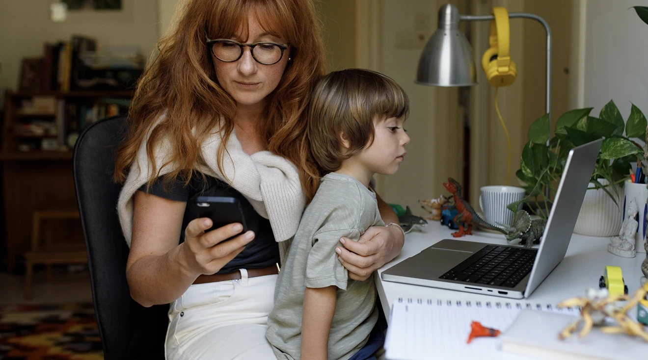 mother sitting at desk with son looking at phone and laptop