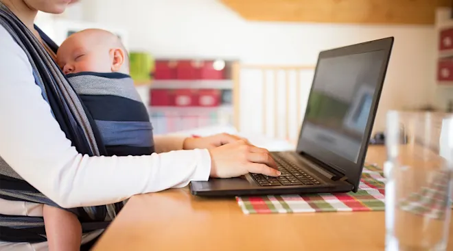 mother wearing baby in wrap while working on laptop at home