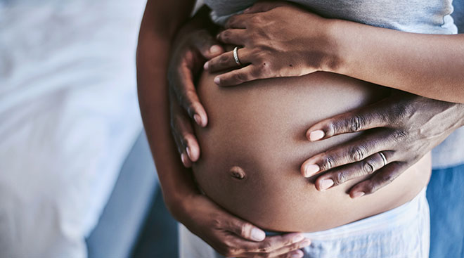 Black pregnant mother cradles her baby bump, with her partner's hands on her belly