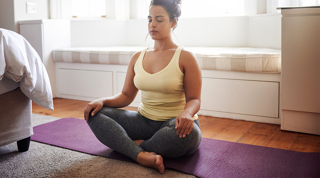 calm woman sitting in her bedroom and doing a breathing exercise on yoga mat