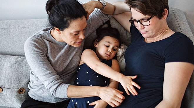 Same sex pregnant couple at home with their daughter on the couch. 