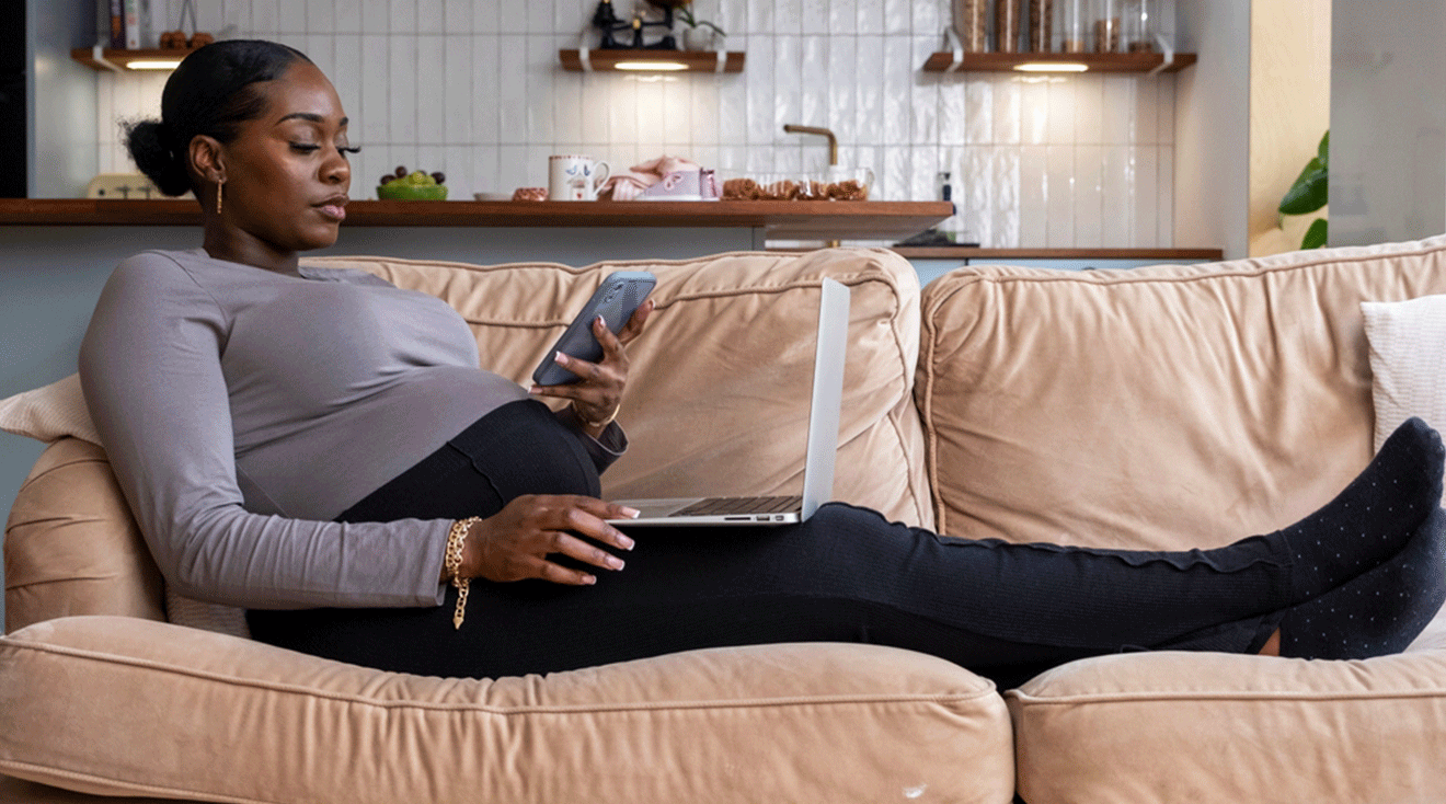 black pregnant woman sitting on couch at home looking at smartphone and laptop