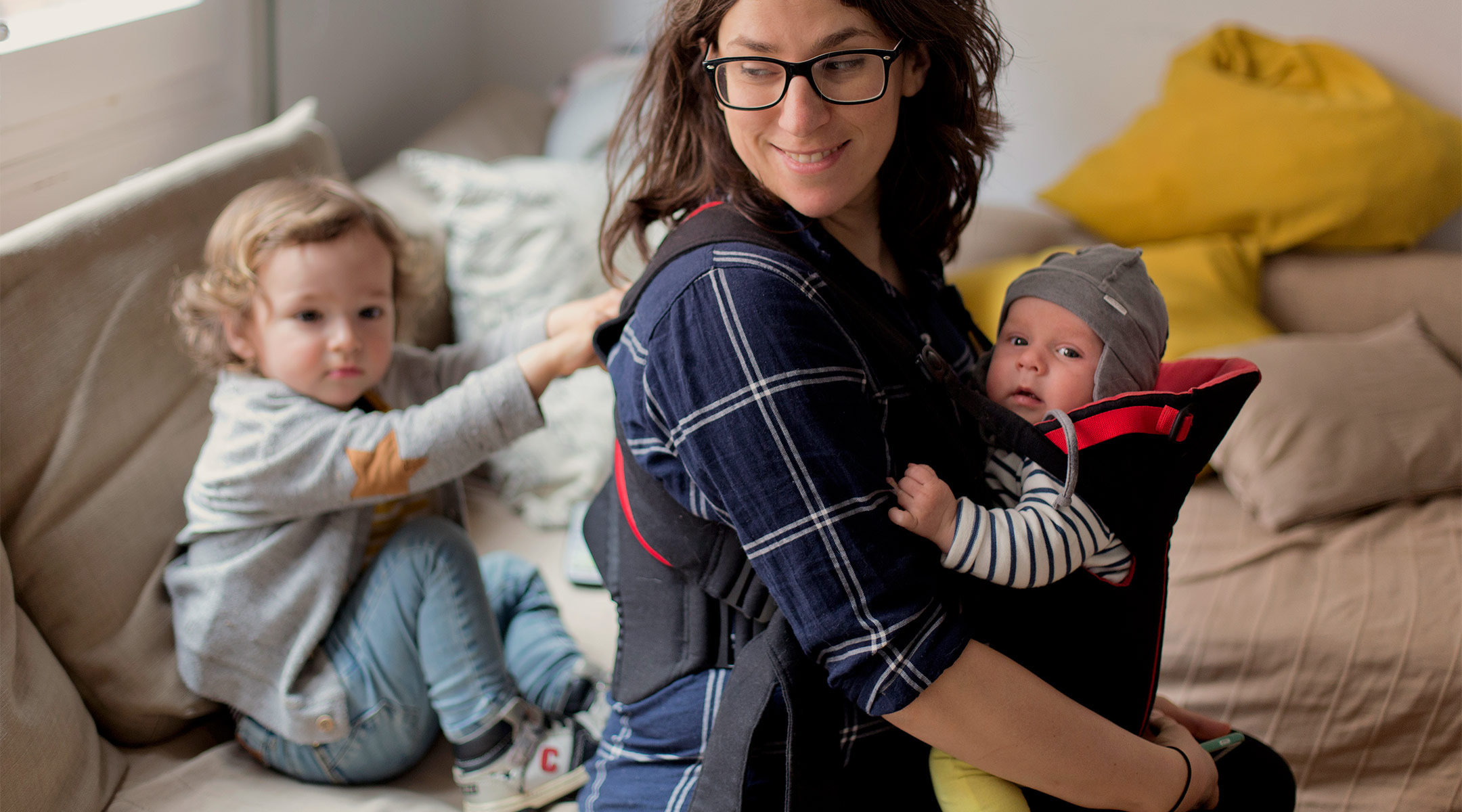 happy mom and home with one child in baby carrier and one tugging at her