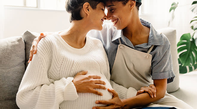 pregnant lesbian couple embracing and smiling on couch at home