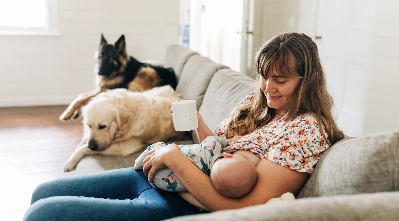 mother drinking a cup of tea while breastfeeding baby on the couch at home