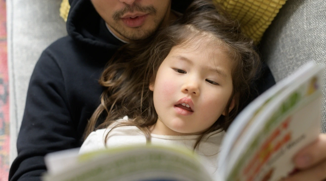 asian father reading book with daughter on couch at home