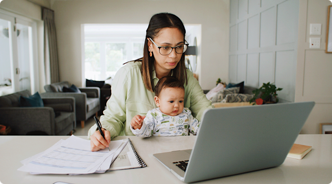 A young mom with her baby on her lap sitting at a table and looking at her laptop.