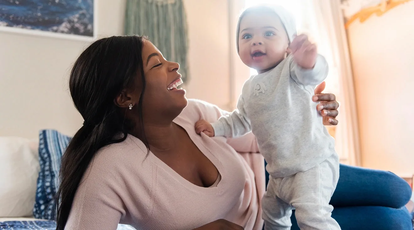 baby standing on bed with smiling mother