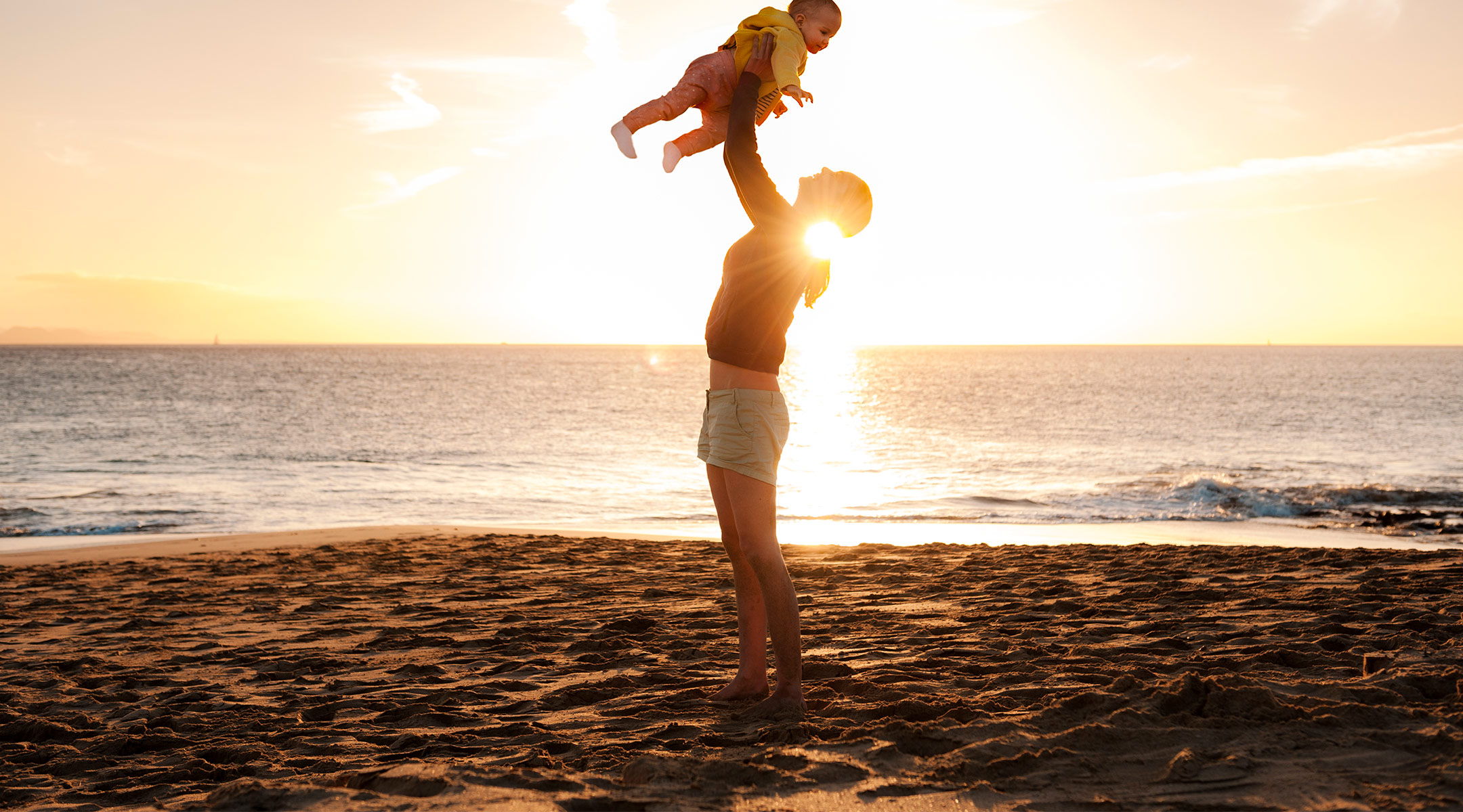 mom traveling with baby, lifting her into the air at the beach