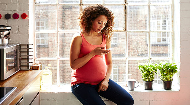 pregnant woman in kitchen by window looking at her phone