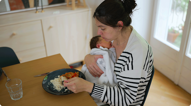 A postpartum mother eating at the kitchen table while holding her newborn baby.