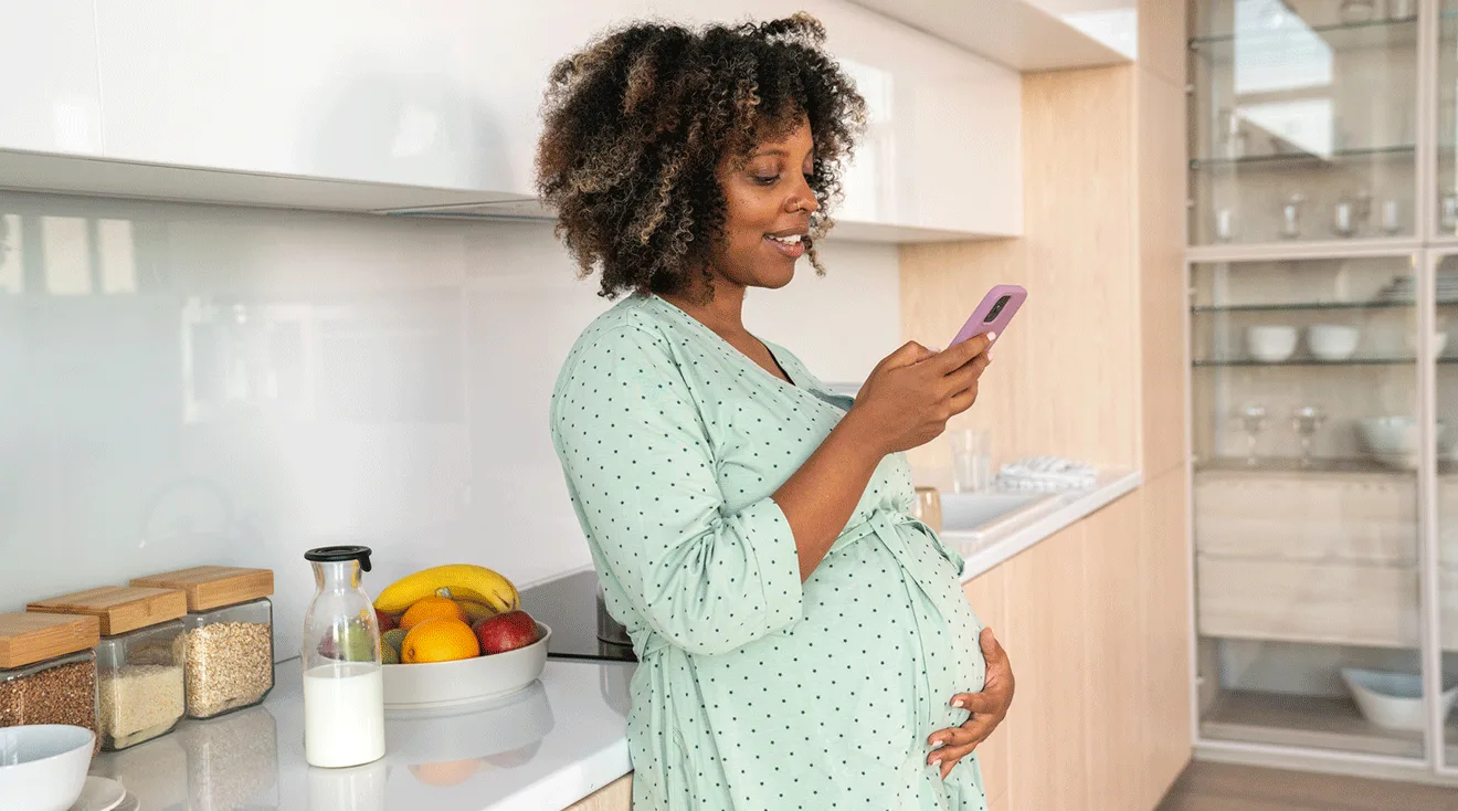 smiling pregnant woman looking at smartphone while standing in kitchen at home