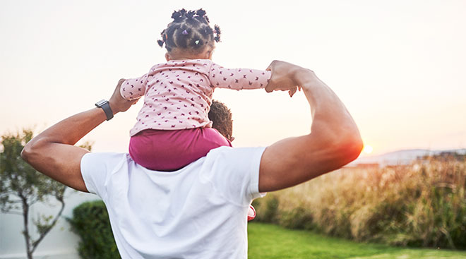 Dad holds his toddler daughter on his shoulders while looking out over an outdoor landscape. 