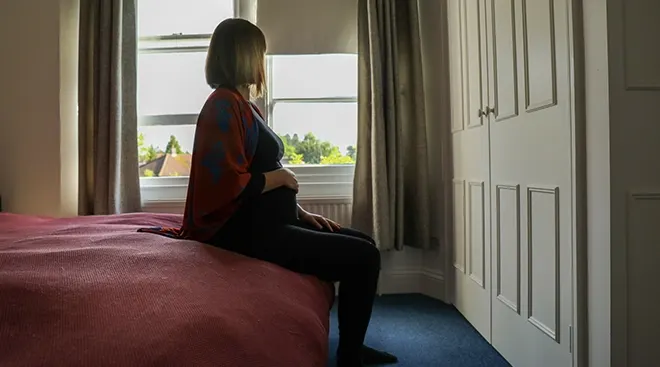 pregnant woman sitting on bed looking out bedroom window
