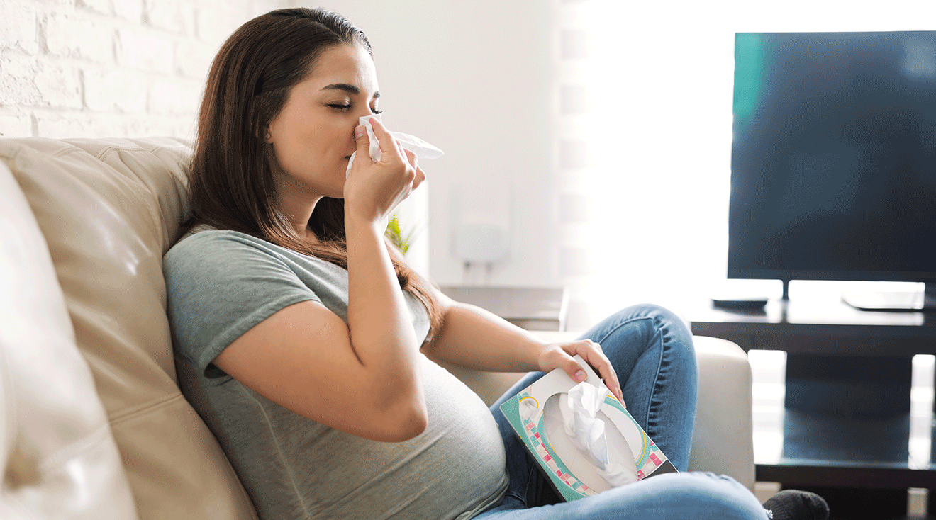 congested pregnant woman blowing nose while sitting on couch at home