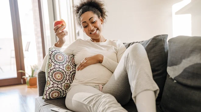 smiling pregnant woman eating apple on couch at home