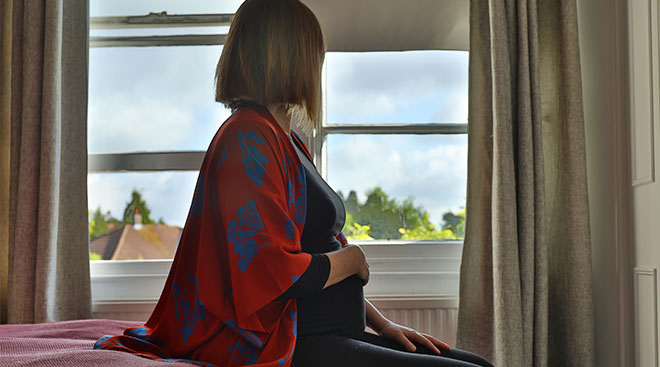 pregnant woman in serious indoor setting sitting on bed and looking out the window