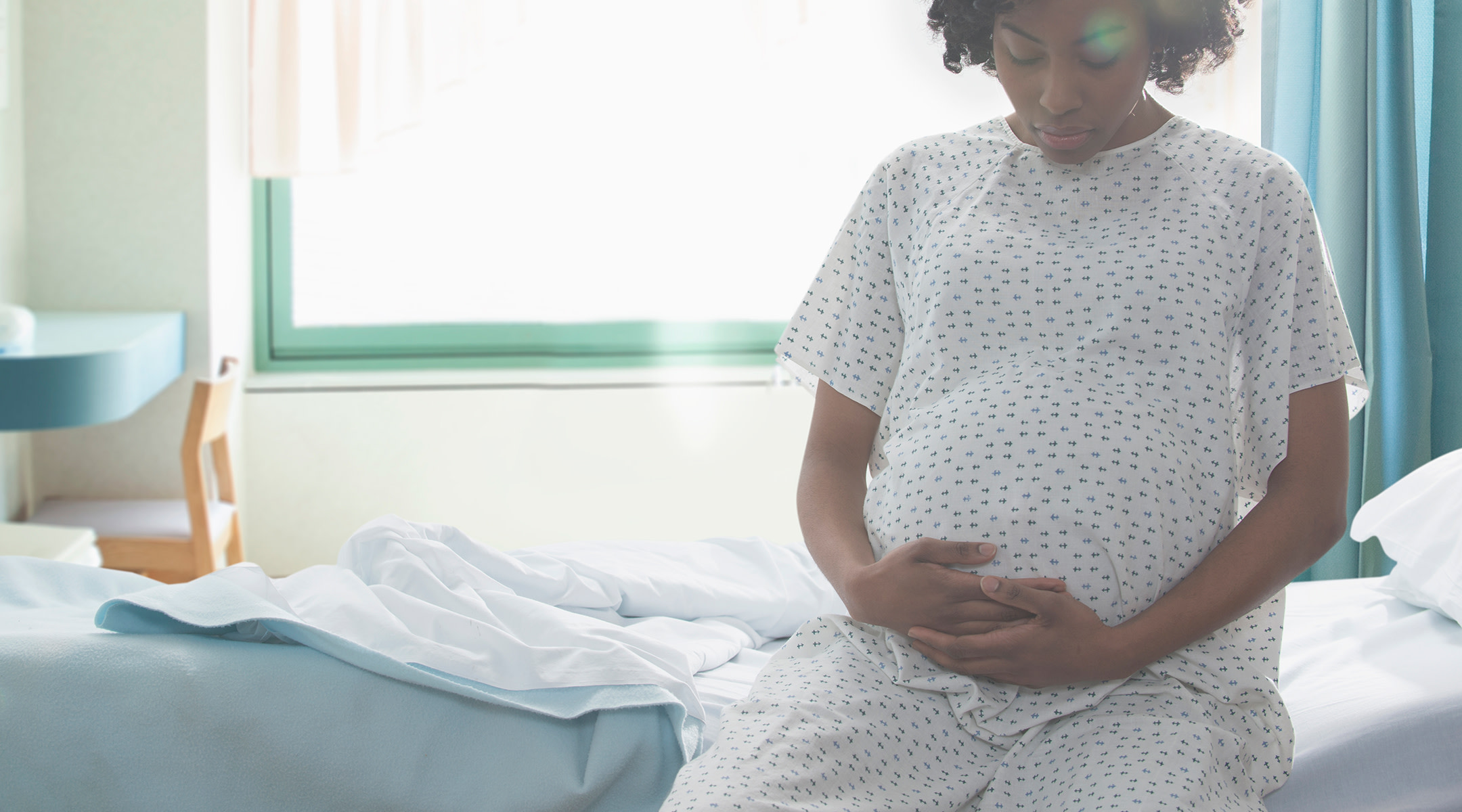 Pregnant mom holding belly in hospital gown, bed; UK mom and baby have lifesaving surgery from inside womb 